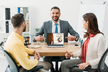 advisor pointing with fingers at laptop with tickets online website on screen wile man and woman thumbing up in office