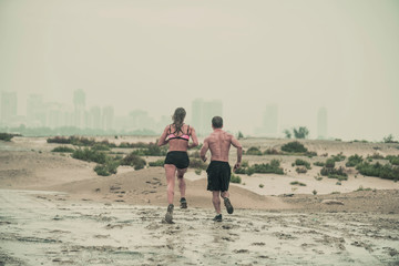 Rear view of muscular male and female athlete covered in mud running down a rough terrain with a desert background in an extreme sport race with grungy textured finish