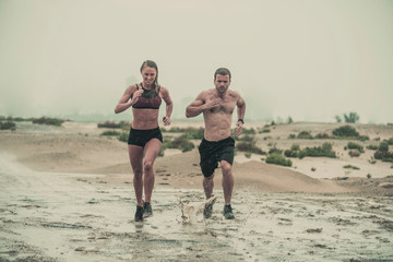 Muscular male and female athlete covered in mud running down a rough terrain with a desert background in an extreme sport race with grungy textured finish