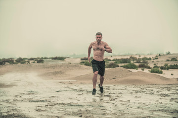 Muscular male  athlete covered in mud running down a rough terrain with a desert background in an extreme sport race with grungy textured finish