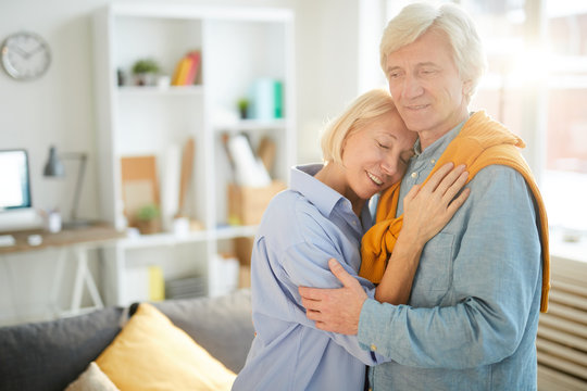 Waist up portrait of happy senior couple embracing tenderly at home lit by sunlight