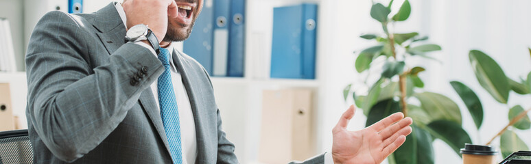cropped view of angry businessman sitting at table with laptop and talking on smartphone in office