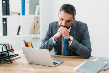 Naklejka premium handsome businessman sitting at table with laptop and thinking in office