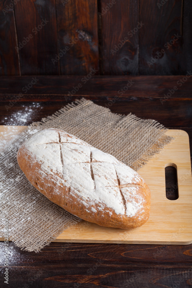 Wall mural fresh brown bread on an oak board with scattered flour on a wooden background with a piece of linen 