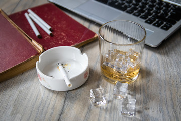 Cigarette in ashtray, glass of whiskey with ice, ice cubes, old book and laptop on wooden table