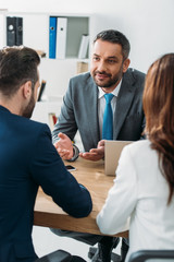 selective focus of handsome advisor in suit talking with investors at workspace
