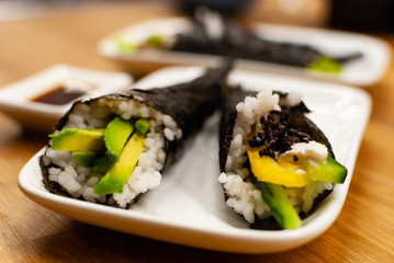 Homemade vegan Temaki-Sushi filled with avocado on a small plate on a wooden table with soy sauce and wasabi