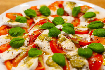 Close up of plate full of vegan Mozzarella made of cashew nuts with tomato slices, basil and pesto on a wooden table