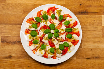 Top view of plate full of vegan Mozzarella made of cashew nuts with tomato slices, basil and pesto on a wooden table