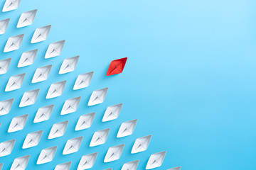 Leadership concept with red paper ship standing out from the group of white paper ships on blue...
