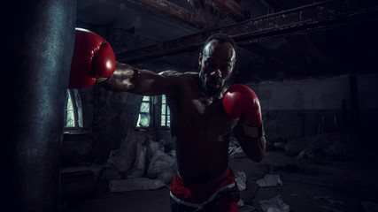 Hands of boxer over dark gym background. Strength, attack and motion concept. Fit african american model in movement. Naked muscular athlete in red gloves. Sporty man during boxing