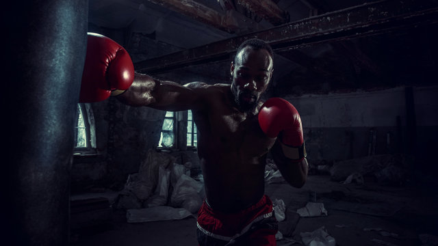 Hands of boxer over dark gym background. Strength, attack and motion concept. Fit african american model in movement. Naked muscular athlete in red gloves. Sporty man during boxing - Powered by Adobe