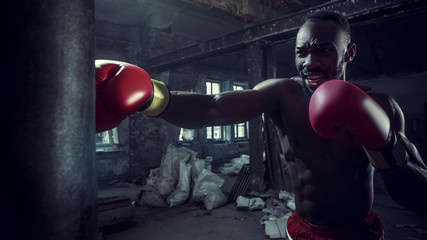 Hands of boxer over dark gym background. Strength, attack and motion concept. Fit african american model in movement. Naked muscular athlete in red gloves. Sporty man during boxing