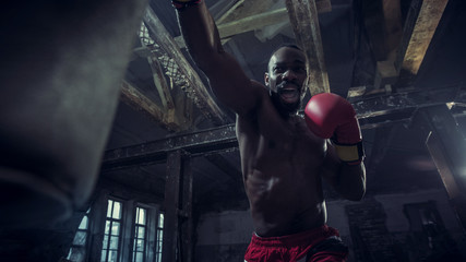 Obraz na płótnie Canvas Hands of boxer over dark gym background. Strength, attack and motion concept. Fit african american model in movement. Naked muscular athlete in red gloves. Sporty man during boxing