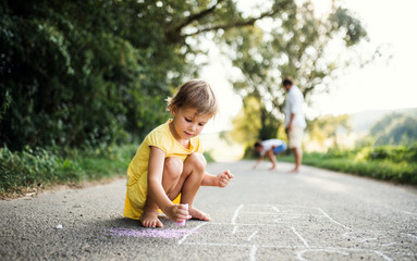 Small cute girl on a road in countryside in sunny summer nature, drawing with chalk.