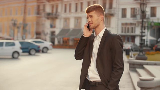 Businessman talking on phone holding hand in pocket at the city square at sunset. Young man in black suite emotionally talking on the cellphone in the street warm morning sun light