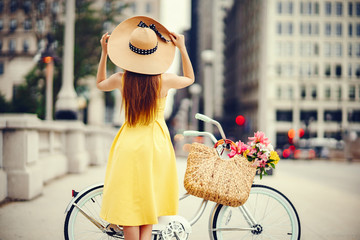 a cute and stylish girl in a yellow dress and long hair rides on a bicycle with a basket of flowers...