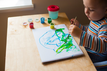 Left hand toddler drawing brush with gouache paint on paper in an album with several colorful...