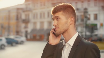 Portrait shot of businessman talking on phone at the city square at sunset. Young man in black suite emotionally talking on the cellphone outdoors in the street warm morning sun light