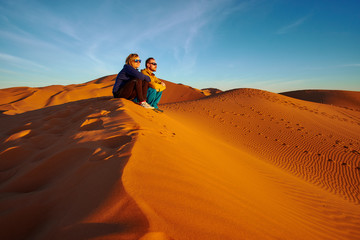 Tourist couple watching sunrise in the desert sitting on a sand dune in Sahara Morocco Africa - obrazy, fototapety, plakaty