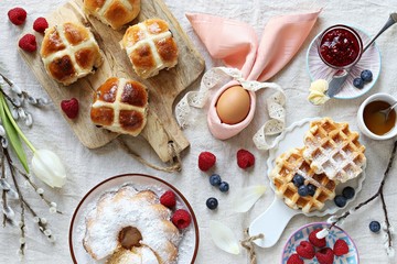 Easter festive dessert table with hot cross buns, cake and waffles on linen table cloth. Overhead...