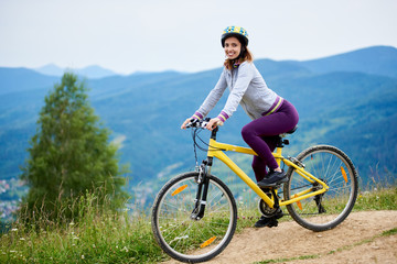 Smiling woman riding on yellow bicycle on a rural trail in the mountains, wearing helmet, on cloudy evening. Mountains, forests on the blurred background. Outdoor sport activity, lifestyle concept