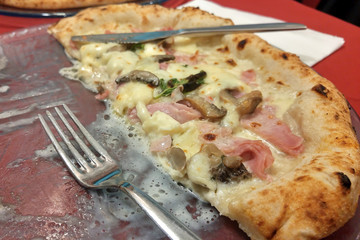 Naples. Half Italian pizza on a plate. Near fork and knife. Juicy pizza with mushrooms, ham and cheese. Seasoned with greens