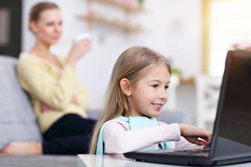 Young mom and her little daughter using laptop and sitting on sofa at home