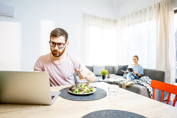 Man working with laptop while eating healthy salad with woman on the background resting on the couch in the modern studio