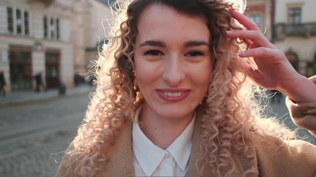 Close-up portrait of beatiful curly hair girl smiling on the camera on the city background. Attractive caucasian woman with curls smiling and looking at camera while standing on street in sunlight.