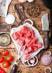 Raw lean diced casserole beef pork steak with vintage meat hatchet and knife and fork on wooden background. Salt and pepper with fresh rosemary, red onion and garlic with rosemary