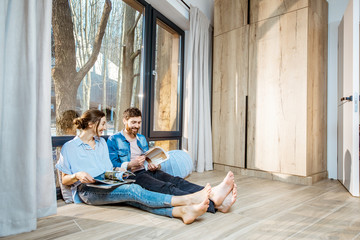 Happy couple sitting together on the floor near the window with beautiful view, reading some magazines and relaxing at home