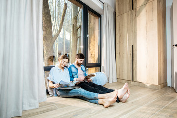 Happy couple sitting together on the floor near the window with beautiful view, reading some magazines and relaxing at home