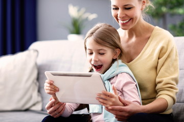 Little girl and her mom using tablet while sitting on sofa at home