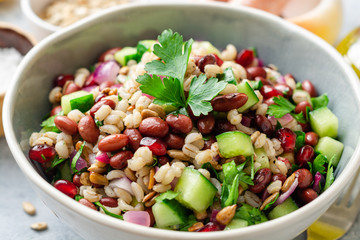 Healthy pearl barley salad with beans, cucumbers, red onion, sunflower seeds, pomegranate and parsley in bowl on concrete background. Selective focus.
