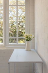 Vertical view of white freesias in jug on table against window and painted brick wall (selective focus)