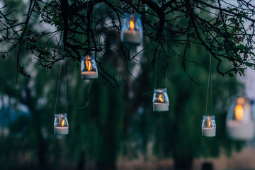 upcycled glass jars garden lanterns illuminated by candles hanging from tree branches in evening - Powered by Adobe