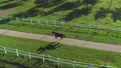 DRONE: Active woman galloping down an empty trail leading through sunlit ranch.