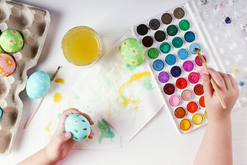 Kid hand painted Easter eggs, paints and brushes on a white table. Preparation for the holiday. Girls hands draw a pattern.