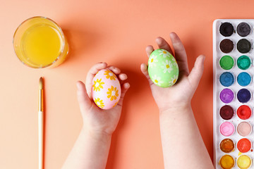 Kid hand painted Easter eggs, paints and brushes on a coral background. Preparation for the holiday. Girls hands draw a pattern.