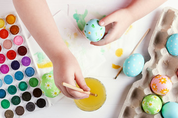 Kid hand painted Easter eggs, paints and brushes on a white table. Preparation for the holiday. Girls hands draw a pattern.