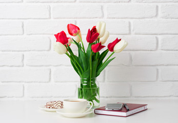 tulip flowers are in a vase on the table, cup of coffee, diary and smartphone, white brick wall as background