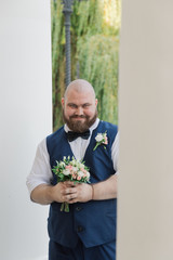 Stylish bearded fat groom with wedding bouquet in the park.