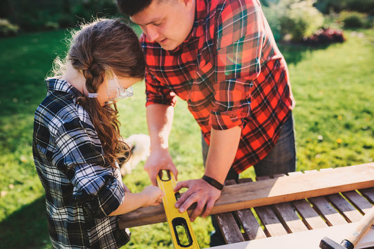 Father Teaching Kid Daughter To Use Tools. Girl Helping Dad With Building Work Outdoor In Summer, Country House On Background