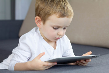 Little blonde boy using touchscreen tablet at home. Happy smart child watching tutorials or playing game on tablet computer. Education, technology, children's security and fun concept.