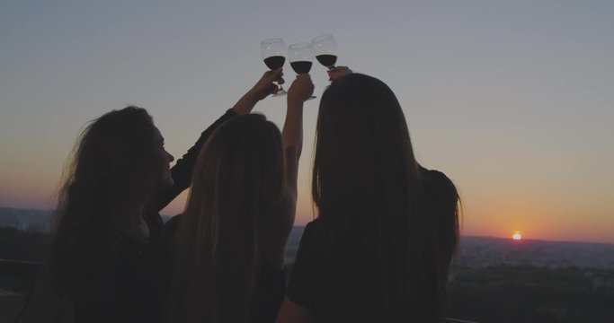 Three ladies at sunset on the loft balcony admire the beautiful sky view , and holding up three glasses of red wine.