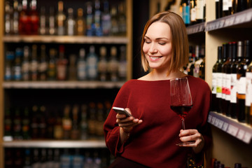 Image of happy blonde with phone and glass in hands at store with wine