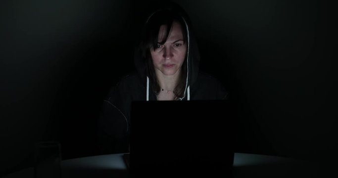 female hacker in a very dark room working on a laptop on the Internet