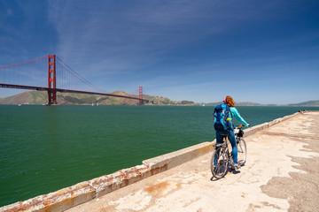 Fototapeta premium Young woman is sitting on a bicycle looking at the Golden Gate bridge from a pier in San Francisco, United States of America