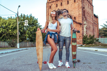 Fototapeta na wymiar Young couple guy with a girl posing on the road in the hands of longboard skates. Happy smiling and sunglasses, summer in the city. Concept sports vacation young family on weekends, trendy hipsters.